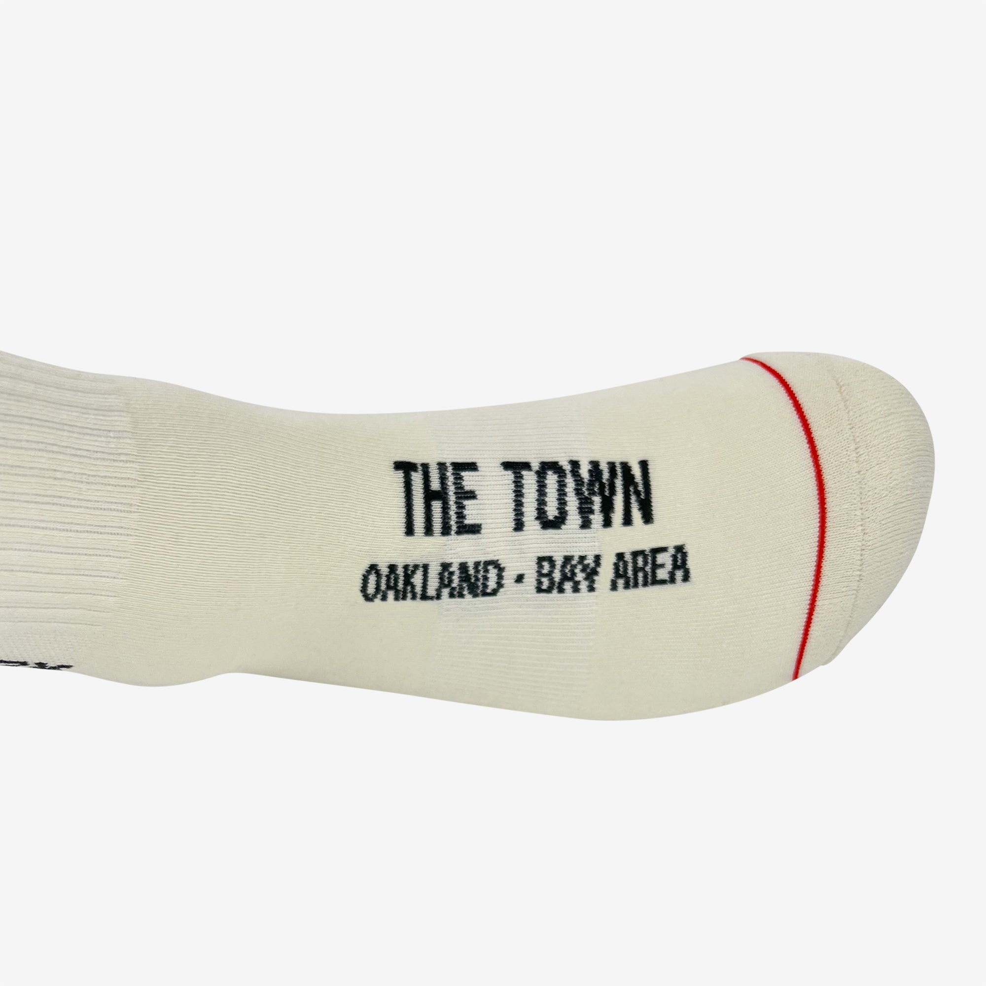 The Oaklandish Utility Sock features a sleek white design with the text "THE TOWN OAKLAND - BAY AREA" in black on the sole and a red trim on the toe area. Constructed from a blend of cotton, polyester, and spandex for optimal comfort and durability, these crew socks are versatile enough to fit most sizes. The socks are displayed laying flat against a plain white background.