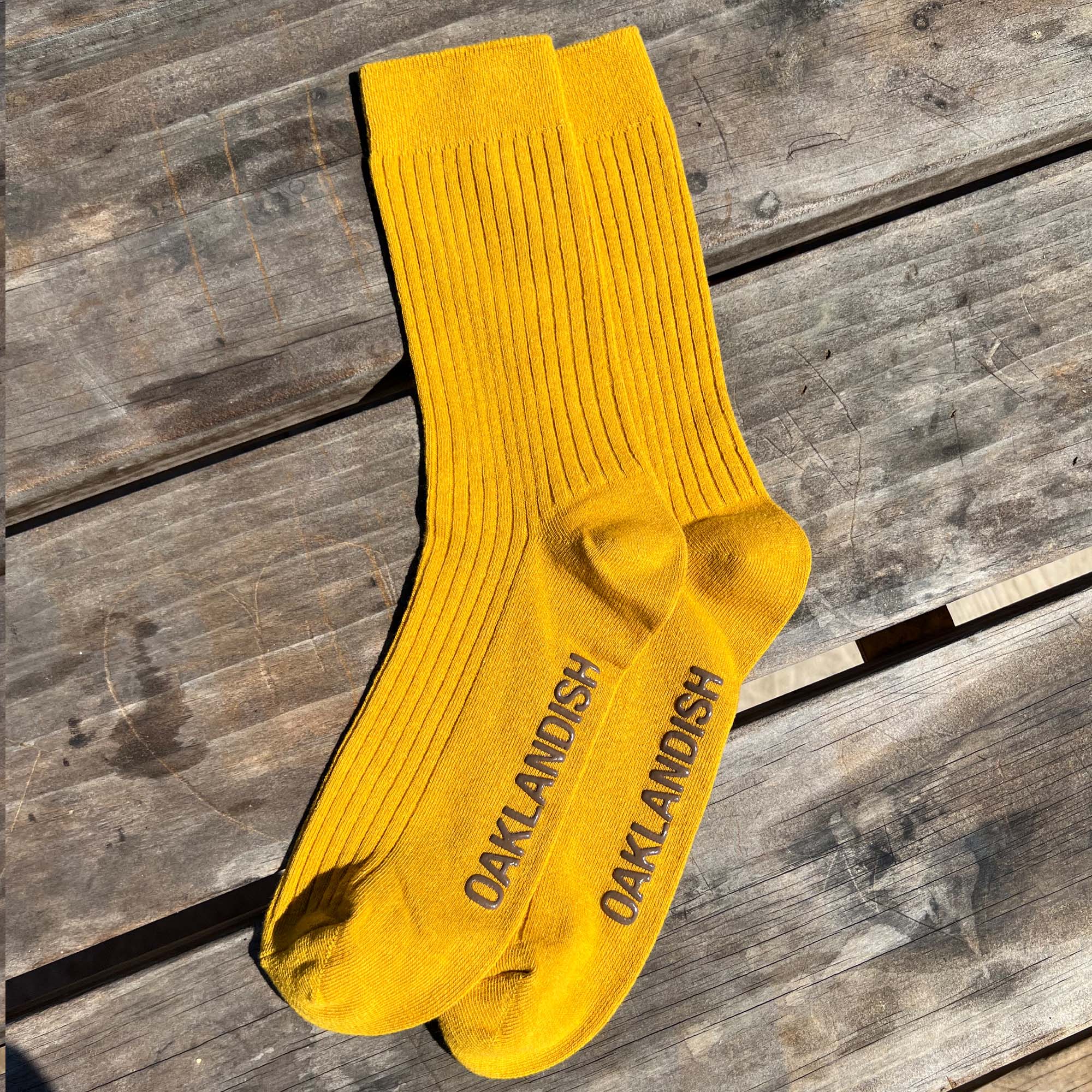 A pair of Oaklandish Grip Crew Socks in vibrant yellow with a ribbed texture are laid out on a weathered wooden surface. "OAKLANDISH" is printed in bold, black letters on the soles. These stylish yellow accessories, made from a blend of cotton, polyester, and spandex, are captured in natural sunlight.