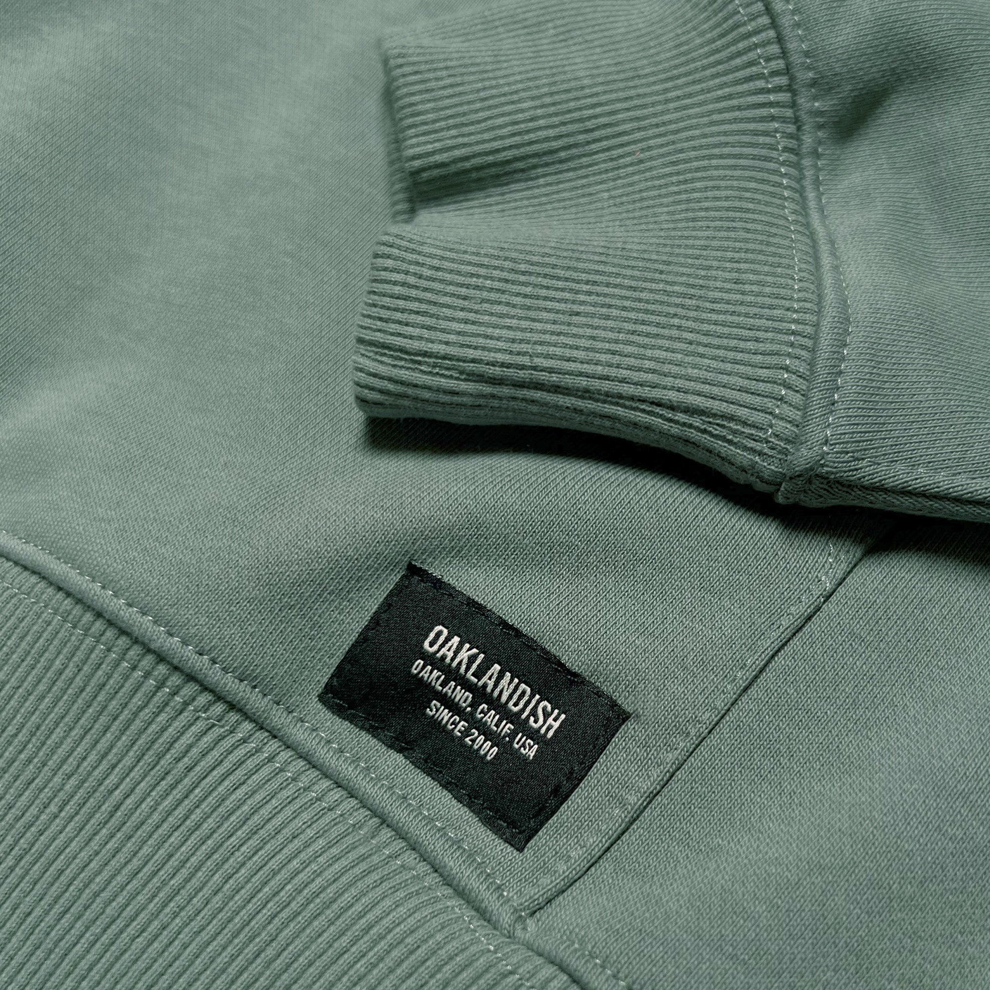 A close-up photograph captures a section of the Oaklandish Premium Hoodie, highlighting the ribbed cuffs and hem of this green heavyweight fleece sweatshirt. Prominently displayed on the fabric is a black sewn label with white text that reads, "OAKLANDISH, Oakland, Calif. USA, Since 2000." The image emphasizes the texture and intricate details of this unisex piece from the brand Oaklandish.