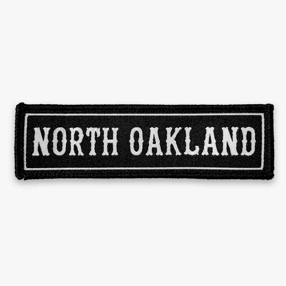 The North Oakland Patch by Oaklandish is a rectangular black iron-on accessory featuring white embroidered text that reads "NORTH OAKLAND" in a bold, old-fashioned font. The text is outlined by a thin white border that follows the edge of the patch, resulting in a clean, minimalist design inspired by Oakland.