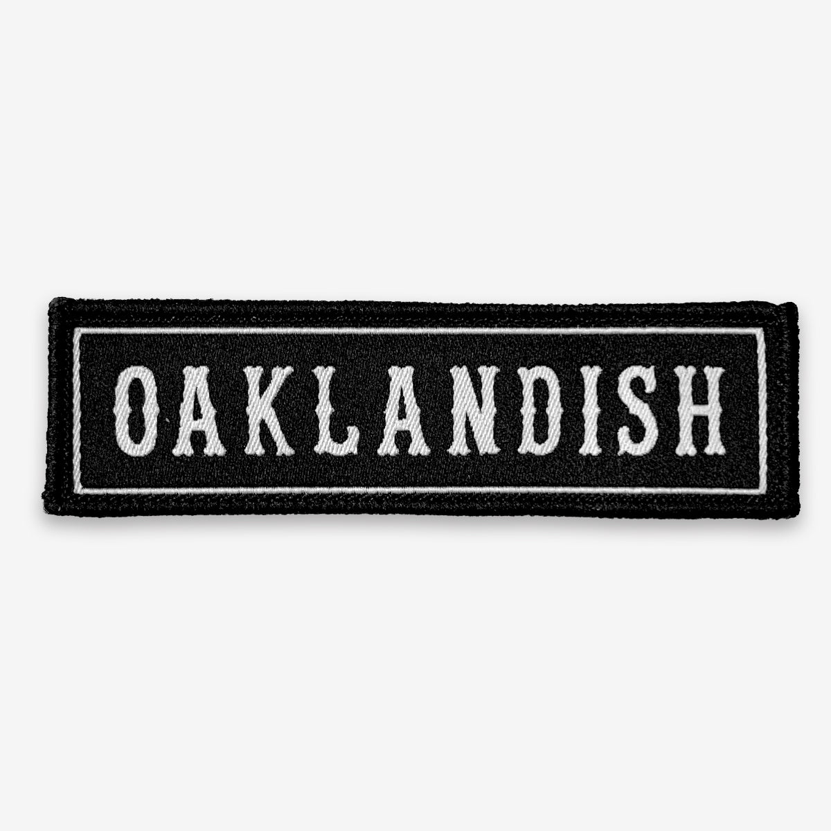 Introducing the **Oaklandish Biker Patch** by **Oaklandish**. This rectangular black patch features a thin white border. Inside the border, the word "OAKLANDISH" is prominently embroidered in all capital letters using a bold, vintage-style font. The simplistic design draws attention to the central text, making it a standout piece against any plain white background. This iron-on biker patch is both stylish and distinctive.