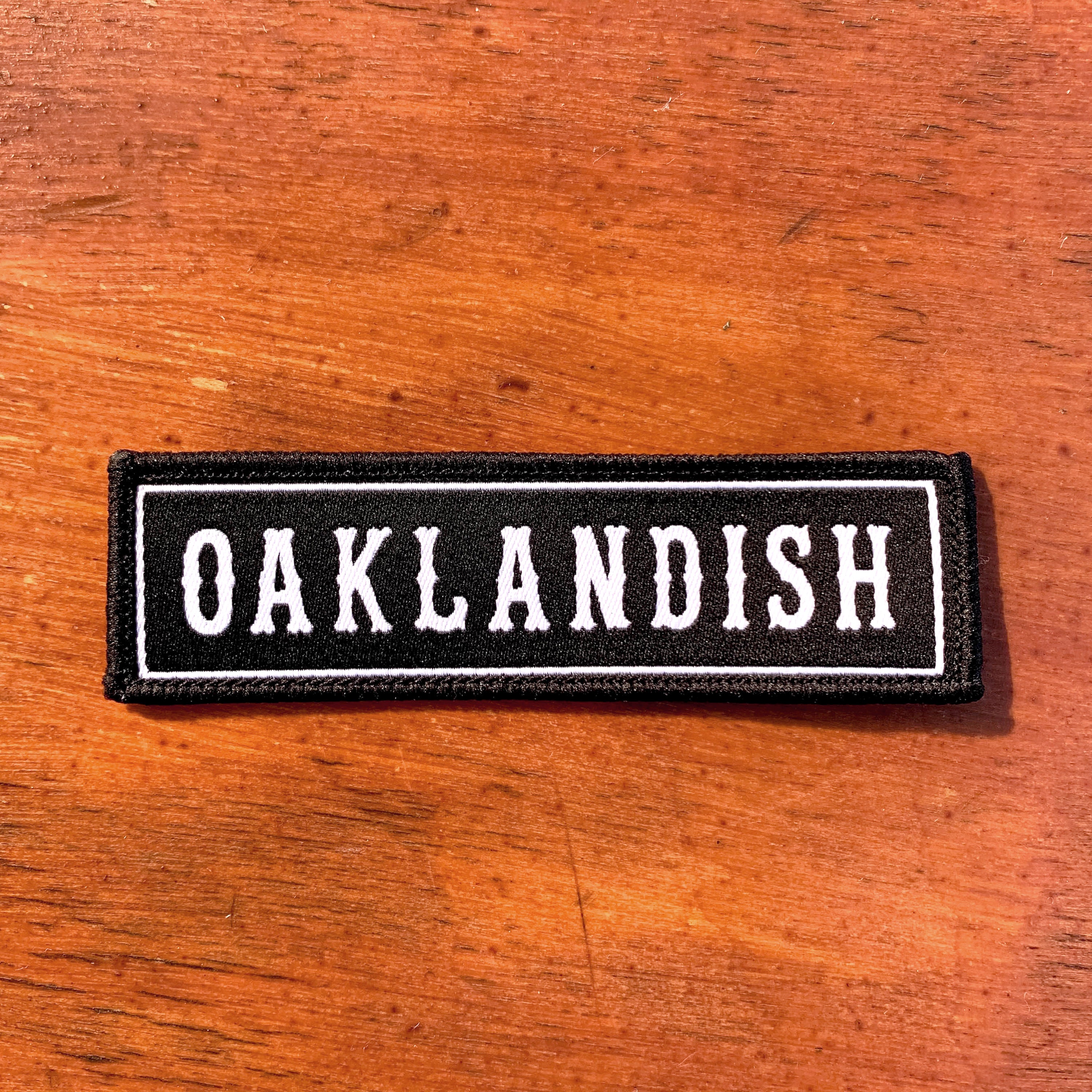 An Oaklandish Biker Patch from the brand Oaklandish features the word "OAKLANDISH" embroidered in white, capital letters on a black background. The iron-on rectangular patch has a white border and is displayed on a brown wooden surface.