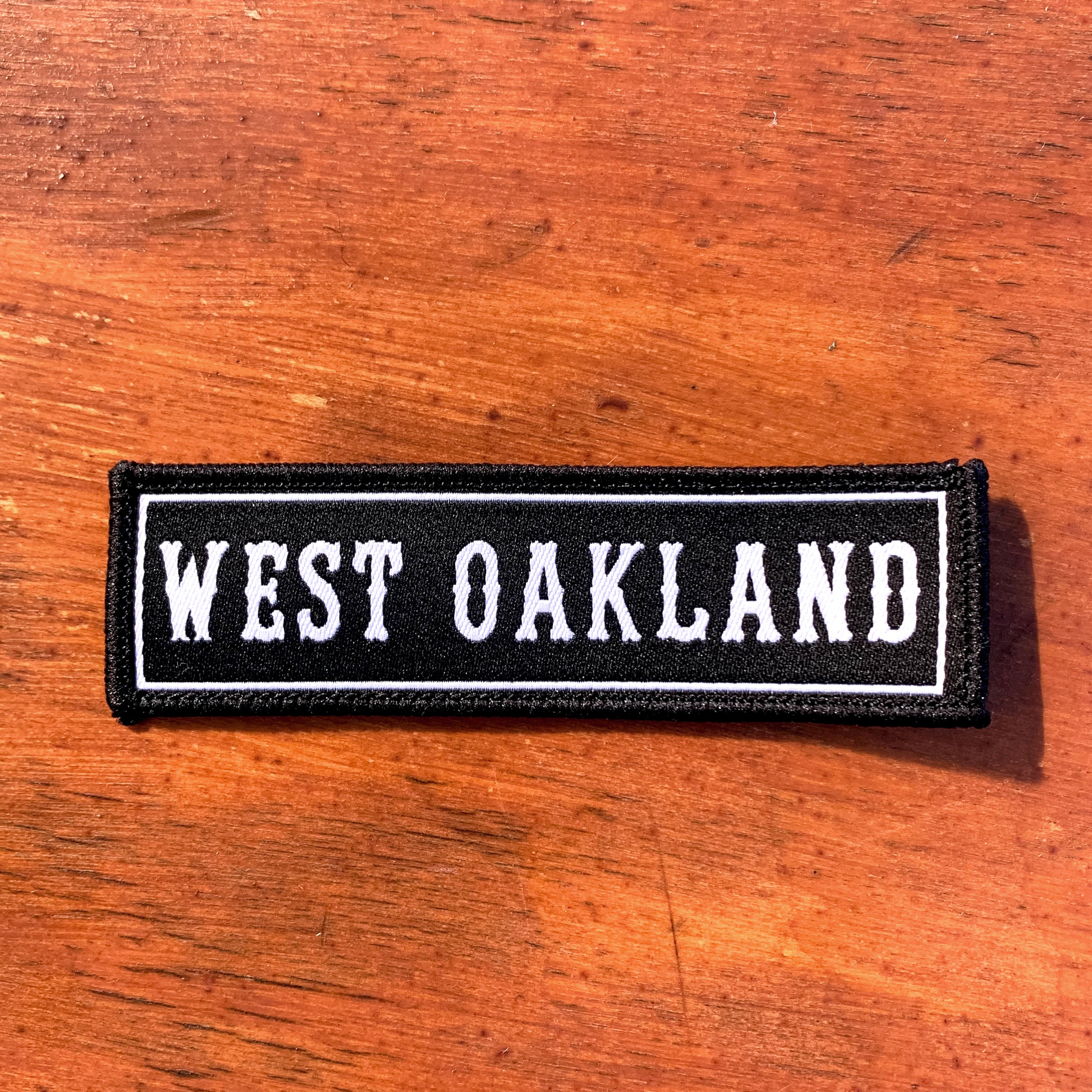 The West Oakland Patch by Oaklandish features a black rectangular base with white embroidered text that reads "WEST OAKLAND" in capital letters. The patch is edged with a thin white border and is displayed on a reddish-brown wooden surface. The slightly distressed, vintage style of the text makes it an ideal accessory for any fan of Oakland.
