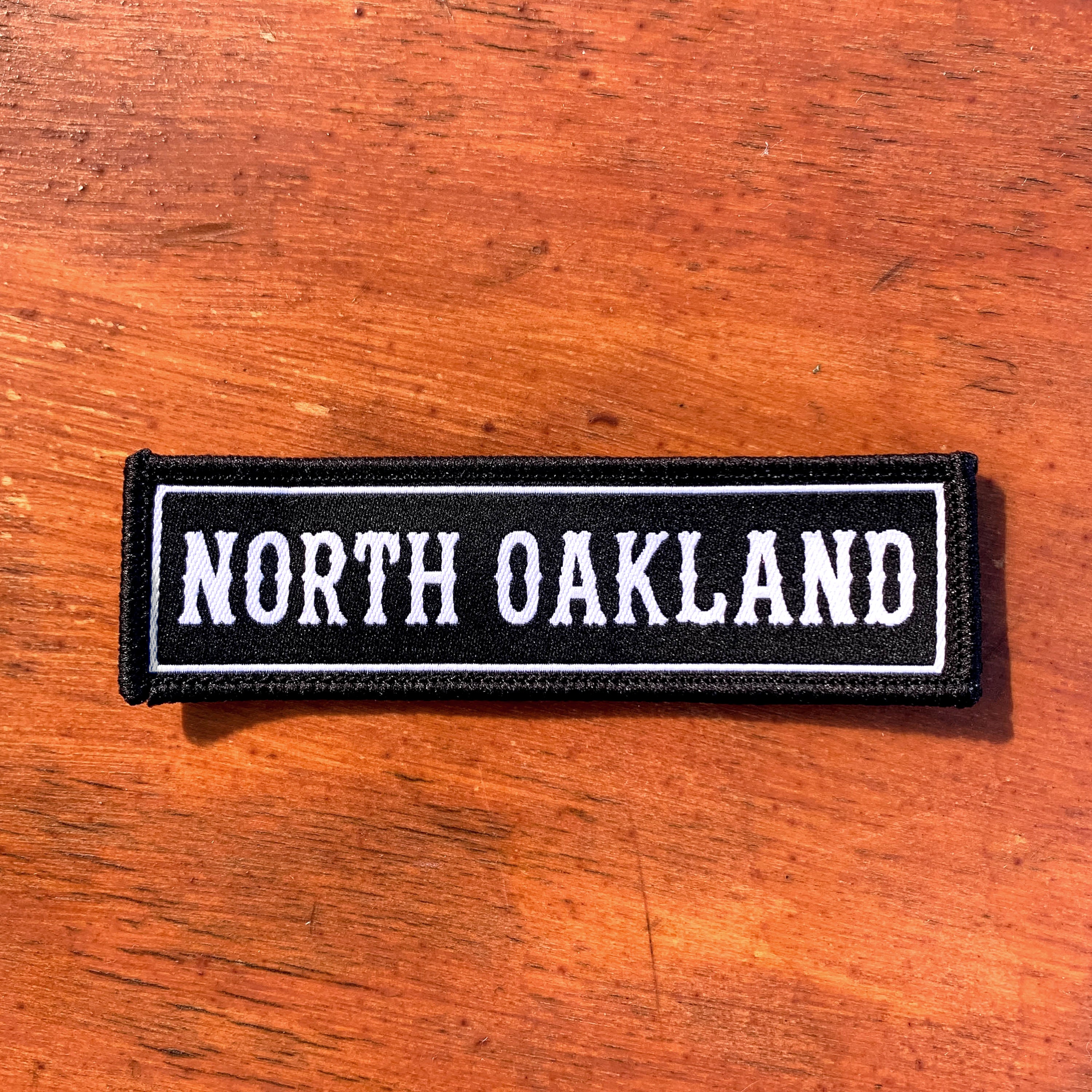 The North Oakland Patch from Oaklandish is a rectangular black iron-on accessory featuring white embroidered text that spells "NORTH OAKLAND" in bold capital letters. It boasts a white border highlighting the text and is beautifully displayed on a wooden surface with a rich, reddish-brown hue and visible grain patterns. This patch is ideal for expressing your Oakland pride.