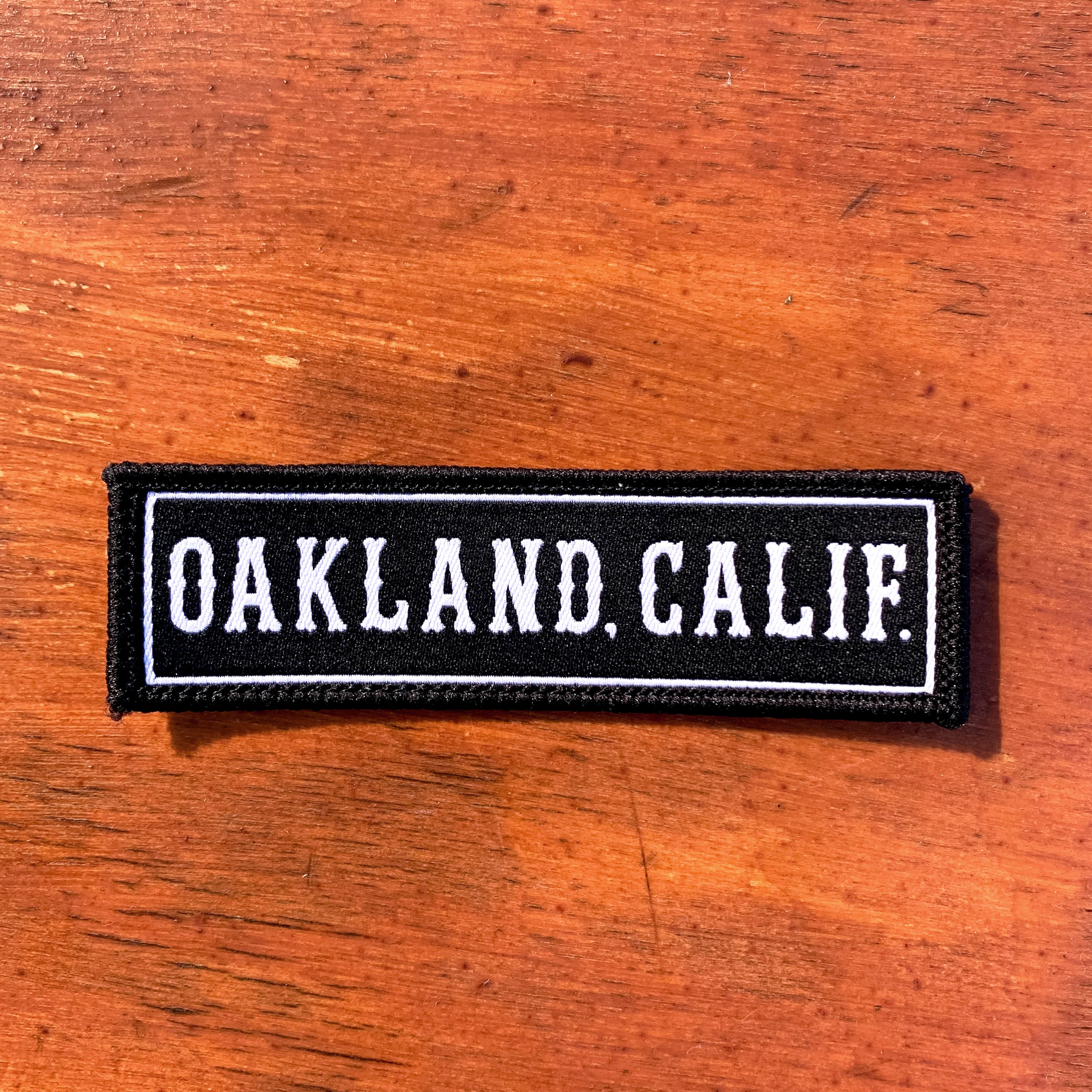 An Oaklandish Oakland CA Patch, black and rectangular, with white embroidery of the text "OAKLAND, CALIF." lies on a polished wooden surface. This iron-on patch features capital letters enclosed within a thin white rectangular border, giving it a crisp, contrasted look against the dark background—pure Oakland vibe.