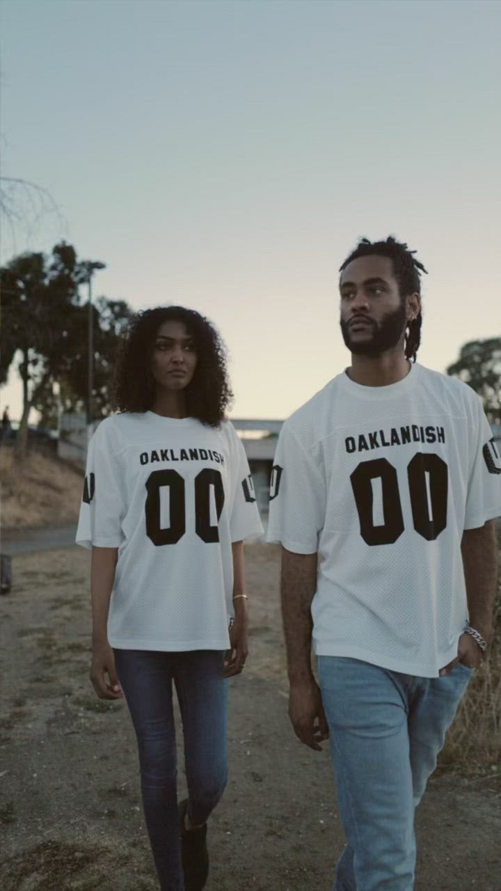 A woman and man standing outside in mesh Oaklandish football jerseys. 