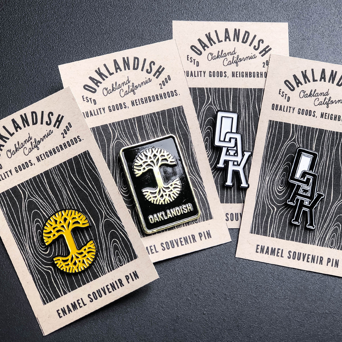 Four enamel lapel pins from "Oaklandish" are displayed. The 1.25" pins feature three distinct designs: two with the Oaklandish tree logo (one in yellow and one in black), and two with the "OAK Monogram." These accessories are attached to rectangular cardboard backings that showcase the "Oaklandish" branding and design details.