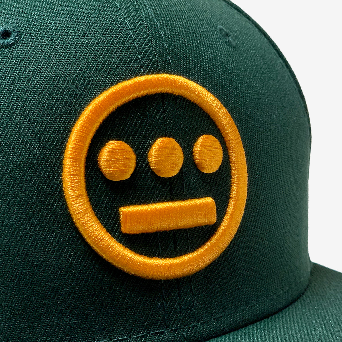 A close-up of a New Era Hiero 59FIFTY Fitted Cap in dark green, showcasing an embroidered yellow hieroglyphics logo. The symbol features a circle with three solid dots evenly spaced along the upper inside edge and one solid horizontal bar centered along the bottom inside edge. The background is plain white.