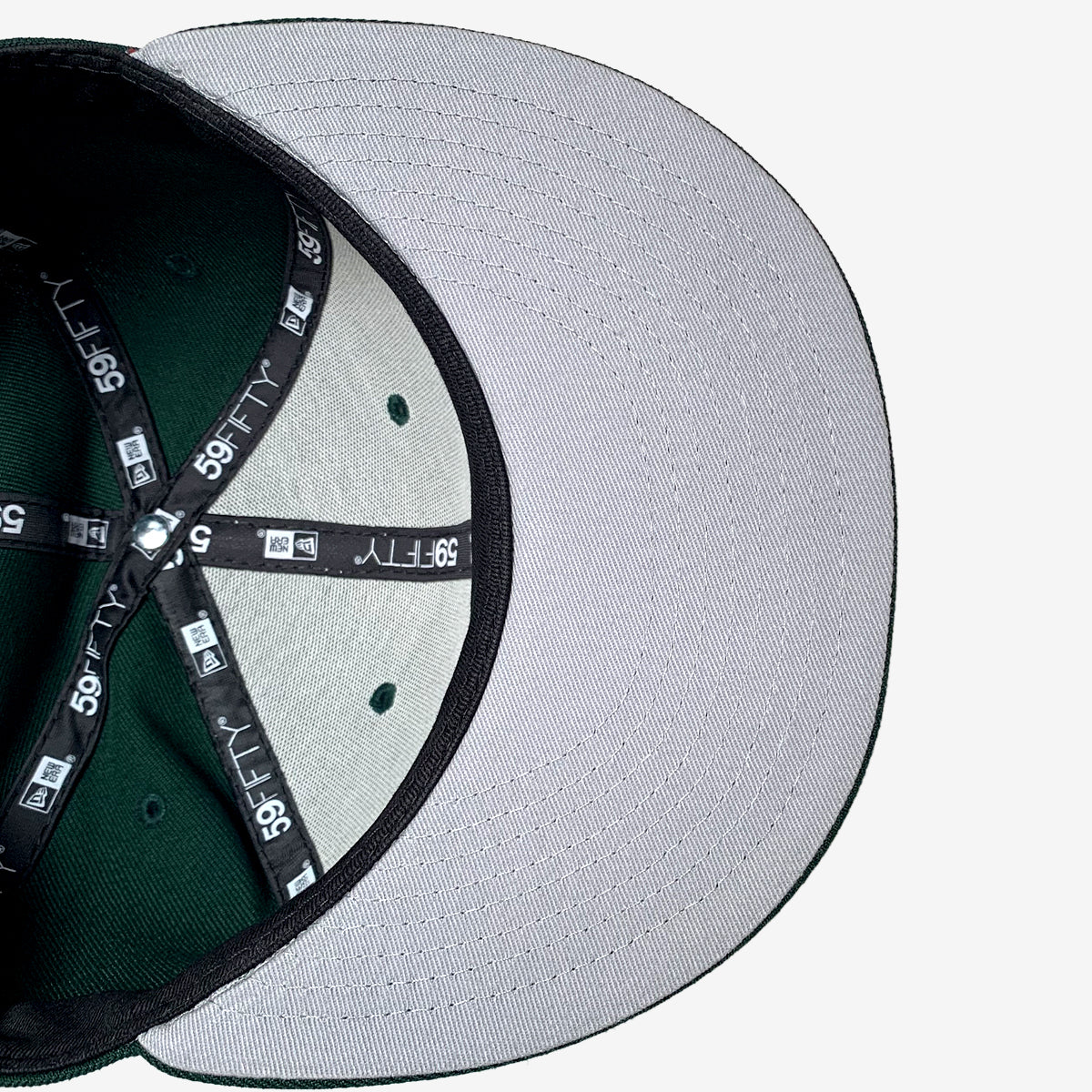 A close-up image of the underside of the New Era Hiero 59FIFTY Fitted Cap by Hieroglyphics. The cap's bill is gray on the underside, and the interior is dark green with black seams. The black inner seams feature white "59FIFTY" text and logos. The hat's size stickers are also visible on the seams.