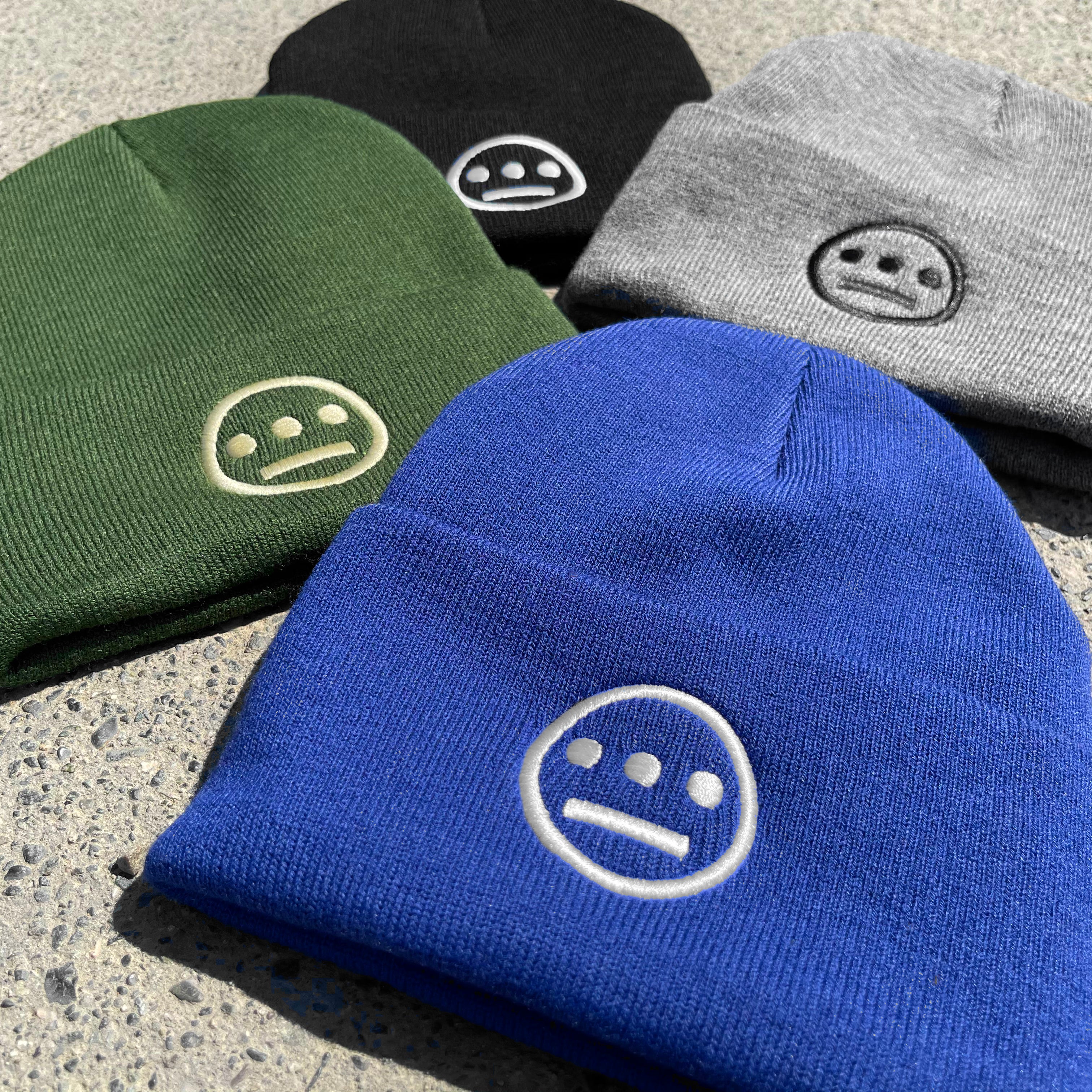 Four Hiero Classic Cuff Beanies, a versatile headwear option from Hieroglyphics, are displayed on a textured surface. The beanies come in black, gray, green, and blue. Each features an embroidered white face with a neutral expression on the front. The designs are arranged neatly, showcasing the variety of colors available.