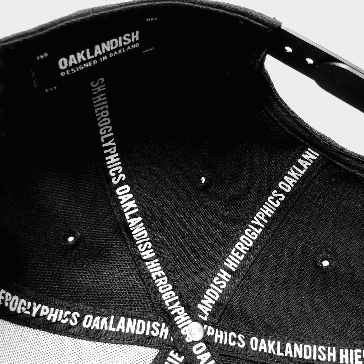 A close-up view of the "Hiero Crew Snapback" cap's interior. The inside labels read "HIERGLYPHICS" repeatedly along the brim and inner seams. "OAKLANDISH DESIGNED IN OAKLAND" is displayed prominently on the inner forehead band. The fabric of this distinctive headwear is attached with metal rivets.