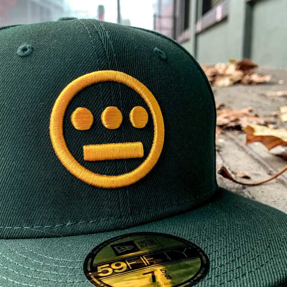 Close-up of the New Era Hiero 59FIFTY Fitted Cap by Hieroglyphics, showcasing its dark green color and flat brim. The cap features a yellow embroidered Hieroglyphics logo with three dots over a horizontal line. It rests on a concrete surface amid scattered autumn leaves, evoking an urban outdoor setting.