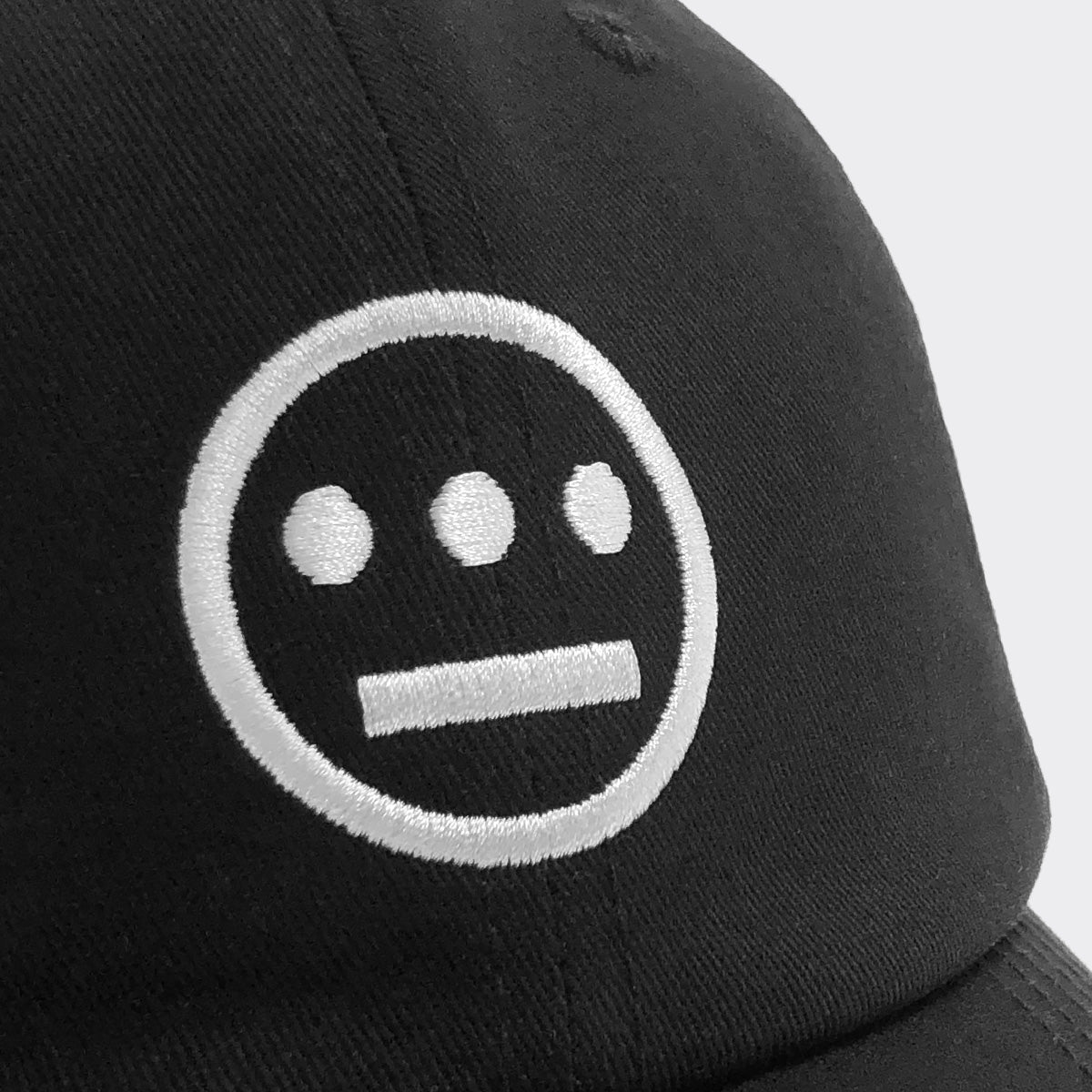 A close-up view of the Hiero Logo Dad Hat from Hieroglyphics features black headwear adorned with an embroidered white logo. The logo depicts a simple, emoticon-like face consisting of three dots for eyes and a horizontal line for a mouth, evoking the style of hieroglyphics. The backdrop is plain gray.
