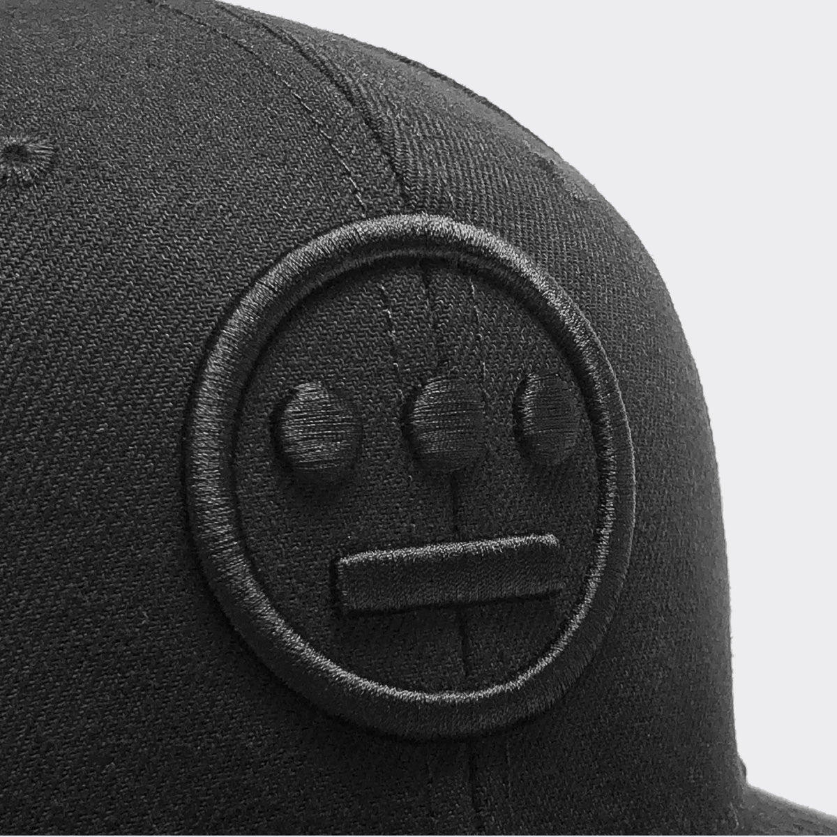 A close-up shot of the Hiero Logo Snapback by Hieroglyphics displays a black baseball cap adorned with an embroidered face design. The face features two large circular eyes and a horizontal line for the mouth, resulting in a neutral expression. This adjustable snapback contrasts strikingly with the plain white background due to its dark color and texture.