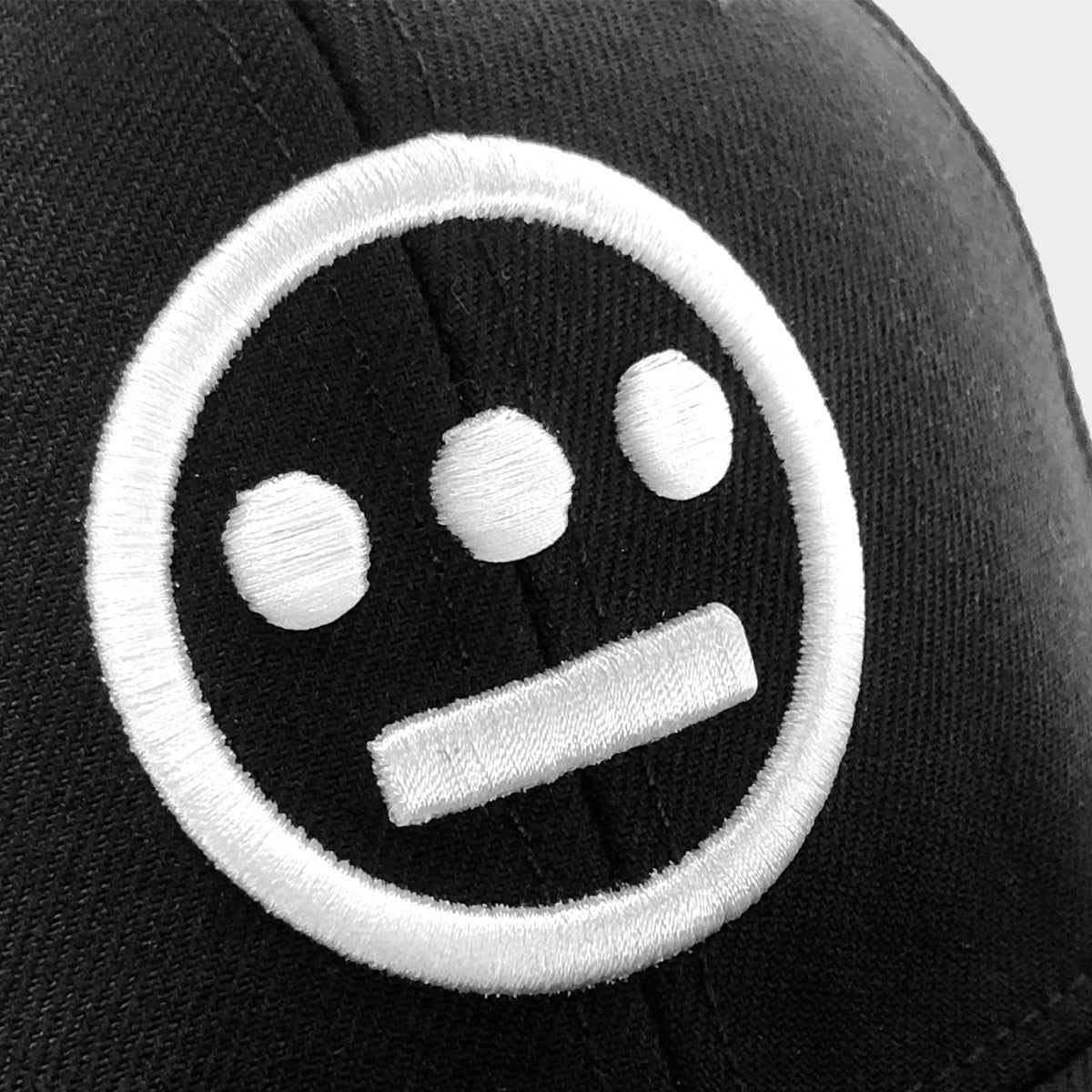 The Hiero Logo Snapback by Hieroglyphics is a black cap with a white embroidered logo. The design features three solid circles in a horizontal line representing eyes and a nose, with a horizontal bar below them as the mouth, all encased in a white circle. This image focuses on the front section of the headwear.