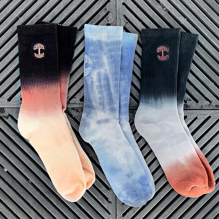 Three pairs of Oaklandish Dawn Fog Socks are showcased against a textured gray surface. From left to right: the first multi-color dip-dyed pair transitions from light pink to black, the middle pair features a blue and white tie-dye pattern, and the third pair fades from gray to dark green with a reddish-brown tip. Each sock has an Oaklandish tree logo near the cuff.