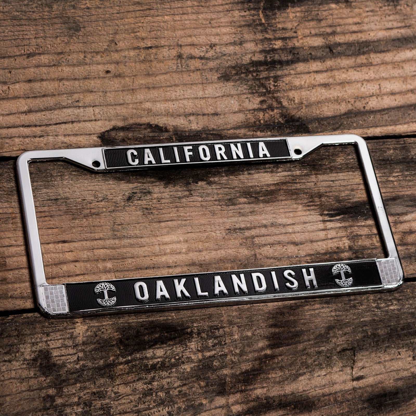 An Oaklandish License Plate Holder, crafted from chromed metal, rests atop a wooden surface. The top part of the frame displays "CALIFORNIA," while "OAKLANDISH" is prominently displayed on the bottom part. Circular emblems with tree designs decorate the lower corners of the frame. This stylish accessory, from the Oaklandish brand, is set against a weathered wood background, enhancing its charm.