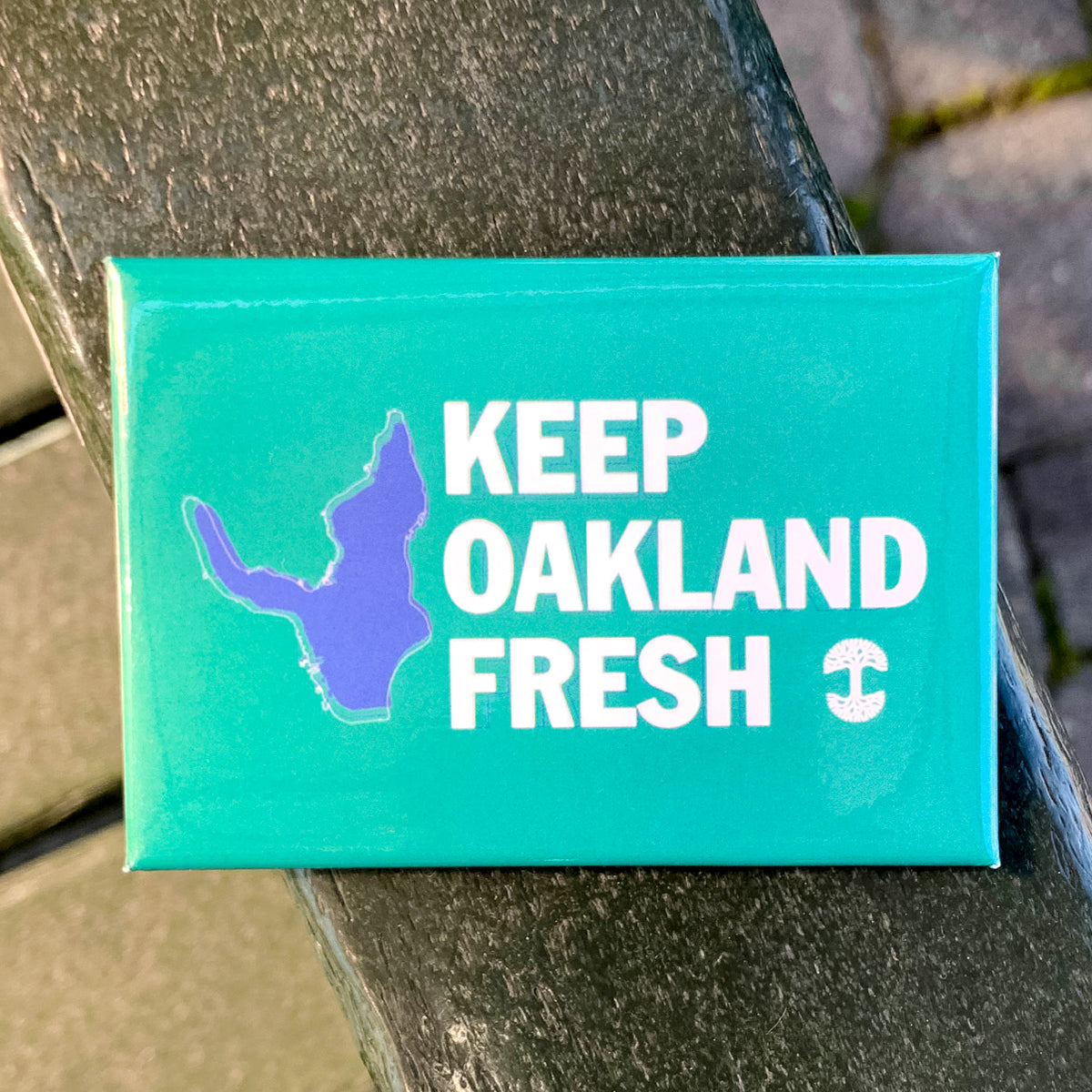 A must-have accessory is the rectangular green "Keep Oakland Fresh Magnet" by Oaklandish, featuring a purple silhouette of Oakland on the left. The white text beside it reads, "KEEP OAKLAND FRESH," with a small stylized tree symbol beneath. The magnet rests on a wooden bench against a blurred backdrop of bricks and greenery.