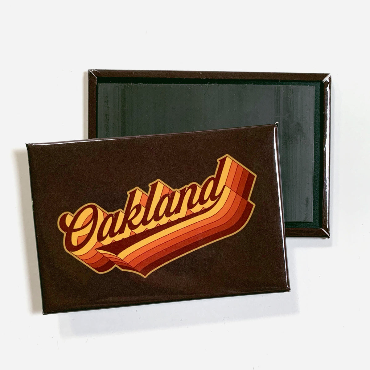 A retro-styled rectangular Swash Magnet by Oaklandish features "Oakland" prominently written in bold cursive letters over an orange and red striped pattern. The brown border adds to its vintage appeal. Another identical magnet lies flat behind it, displaying the plain black magnetic back. The backdrop is white.