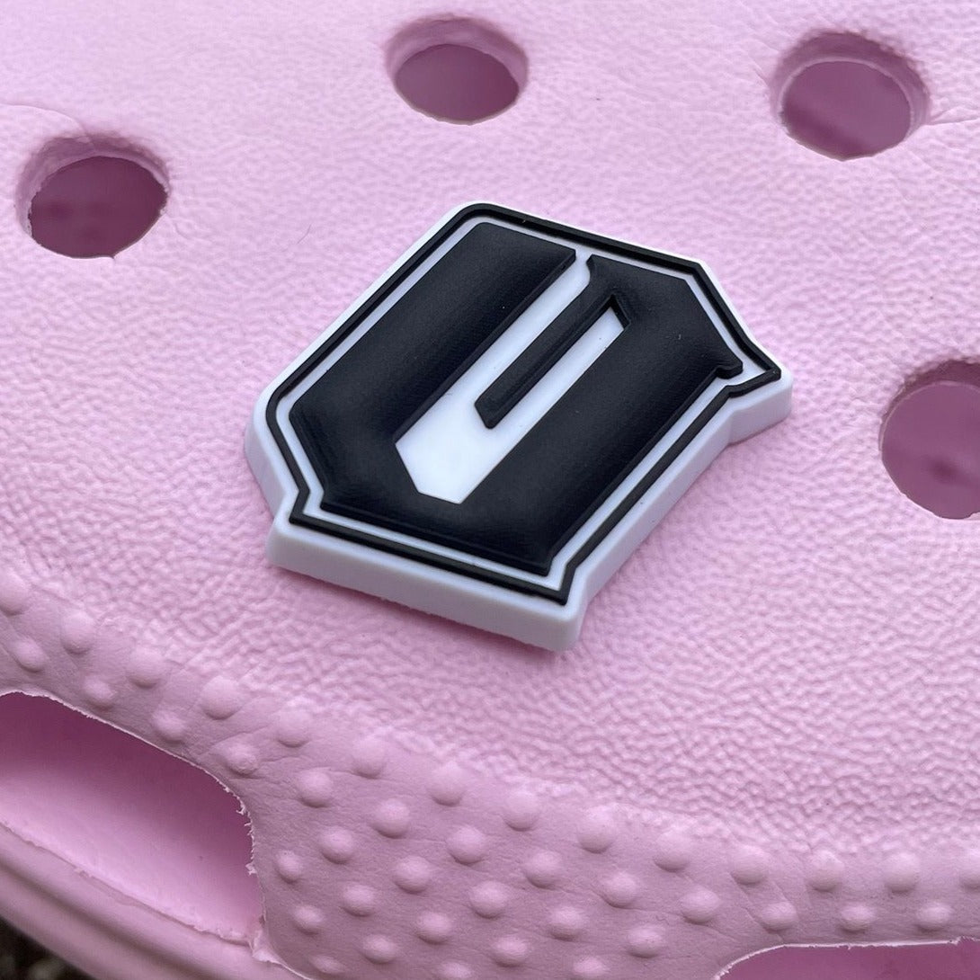 Close-up of a pink foam clog shoe with several holes. An Official Shoe Charm by Oaklandish, featuring a black and white 3D letter "U," is attached to the upper surface, adding a trendy accessory to the design. The bold, sharp-edged charm is positioned centrally within the hole pattern. The shoe also showcases a textured surface.