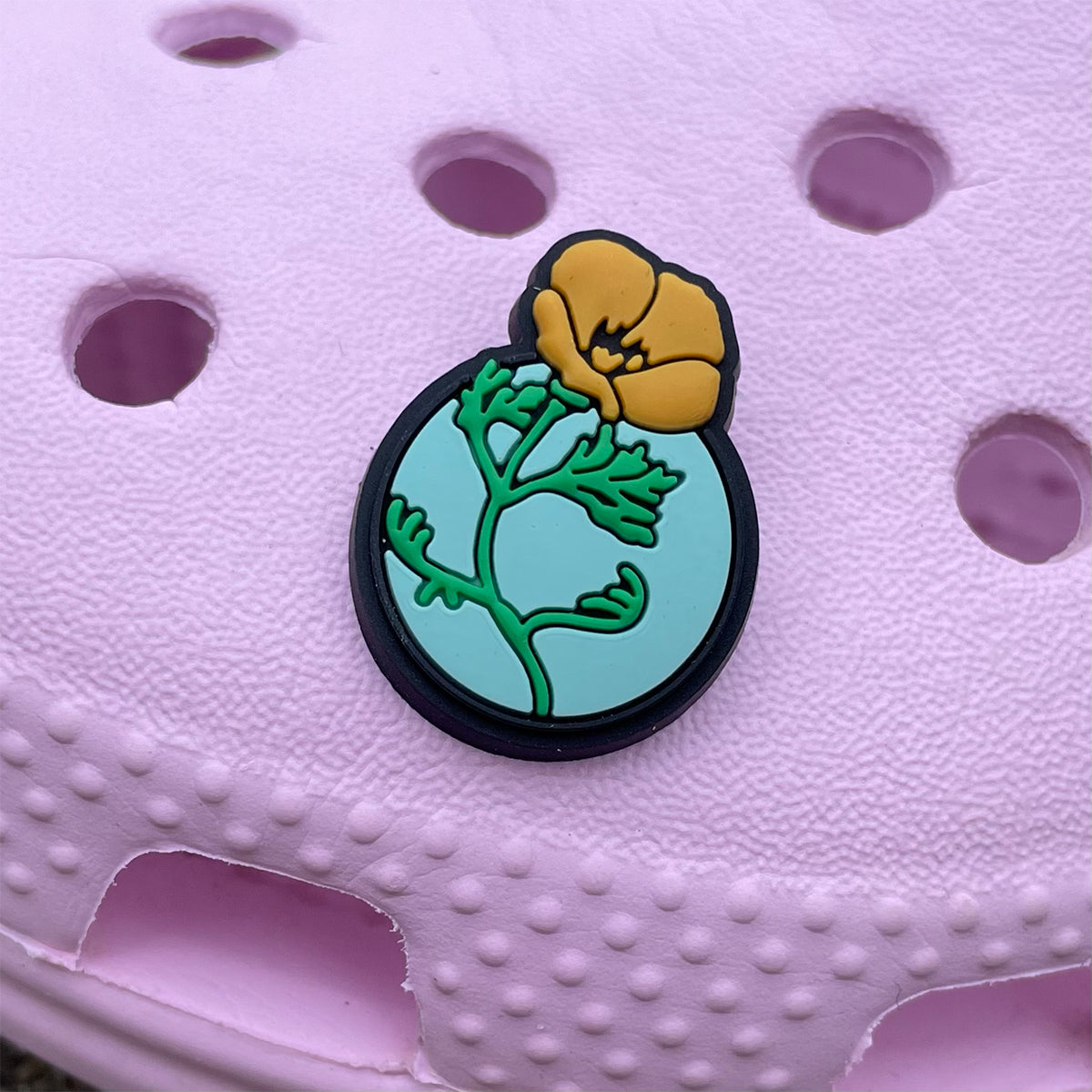 The Oaklandish Poppy Shoe Charm, featuring a vibrant yellow flower with a green stem and leaves, is fastened to a light pink perforated surface with circular and oval holes. The charm boasts a black outline and light blue background that accentuates the striking floral design, adding flair to your clogs.