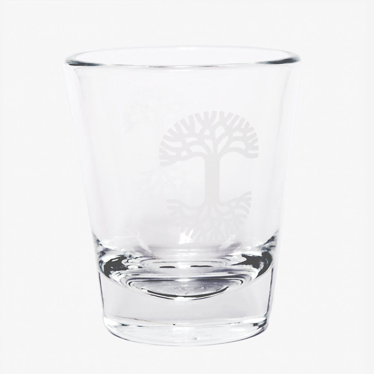 The Oaklandish Trees Shot Glass by Oaklandish is a clear glass tumbler with a thick, solid base, showcasing an elegant logo etching of a stylized tree with sprawling branches and roots, symbolizing the tree of life. This exquisite shot glass is empty and set against a plain white background.