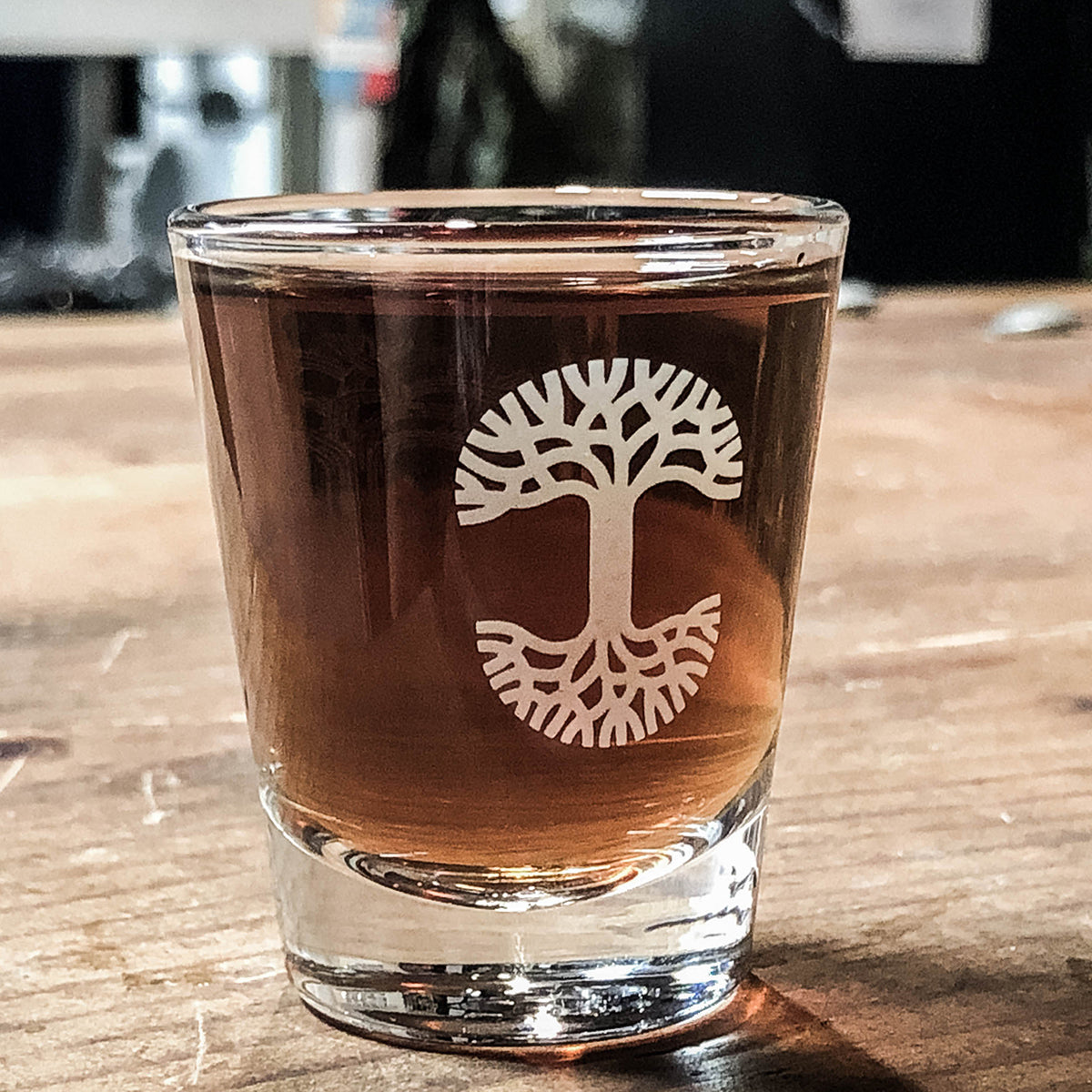 An Oaklandish Trees Shot Glass, filled with an amber-colored liquid, sits on a wooden surface. The clear glass features a distinctive white tree design, resembling an inverted tree with roots spreading out on the bottom half and branches on the upper half. The logo etching draws attention as the background blurs, making the Oaklandish Trees Shot Glass the focal point.