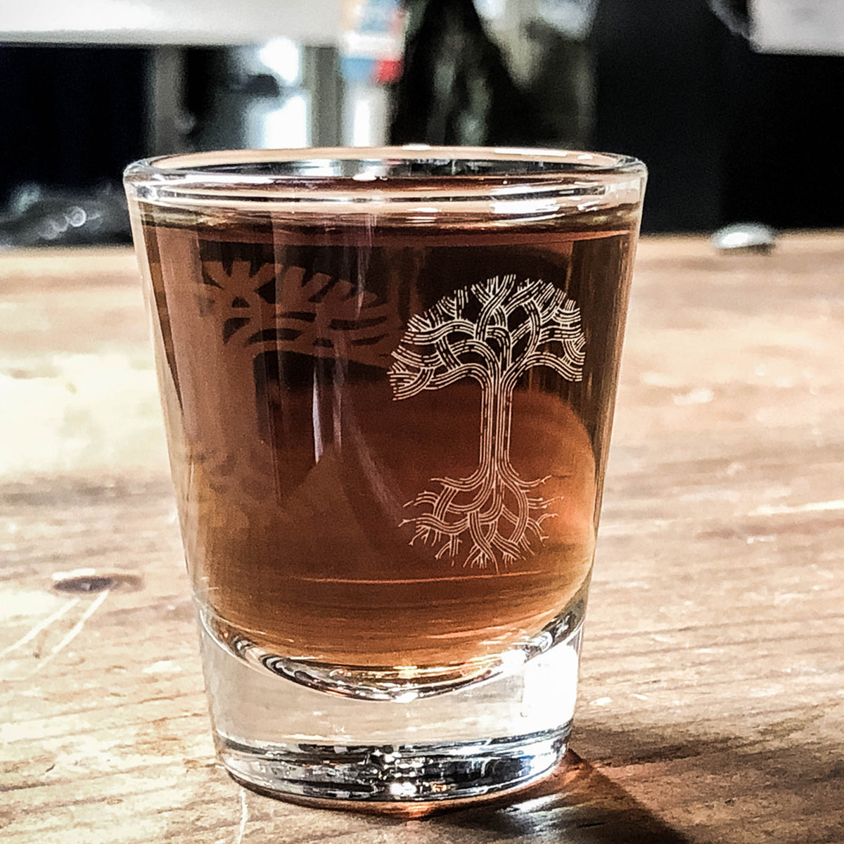 The Oaklandish Trees Shot Glass filled with a brown liquid sits on a rustic wooden surface. The clear glass features an etched tree design with detailed roots and branches. The background is slightly blurred, highlighting the focus on the Oaklandish Trees Shot Glass and its contents.