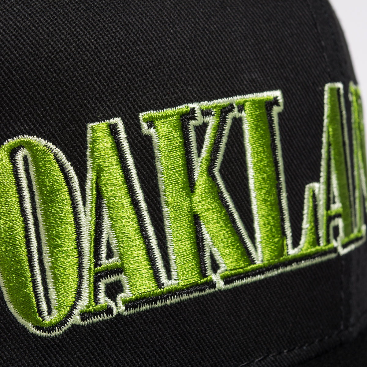 Close-up image of the Greenback Snapback cap from Oaklandish, featuring the word "OAKLAND" embroidered in bold green stitching with white outlines. The black fabric background and centered letters highlight the detailed embroidery work perfectly. Ideal headwear for any Oaklandish fan!