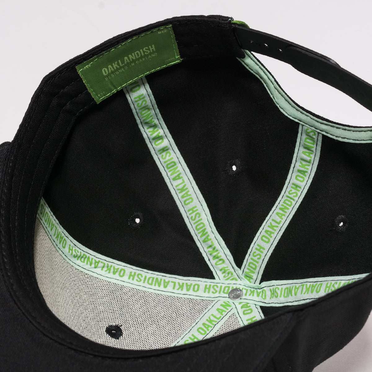 The image showcases the interior of the Oaklandish Greenback Snapback cap. The inner seams and sweatband are lined with green fabric, repeatedly labeled with "OAKLANDISH" in white text. Inside, a green label reads "OAKLANDISH SINCE 2000." Two silver eyelets provide ventilation.