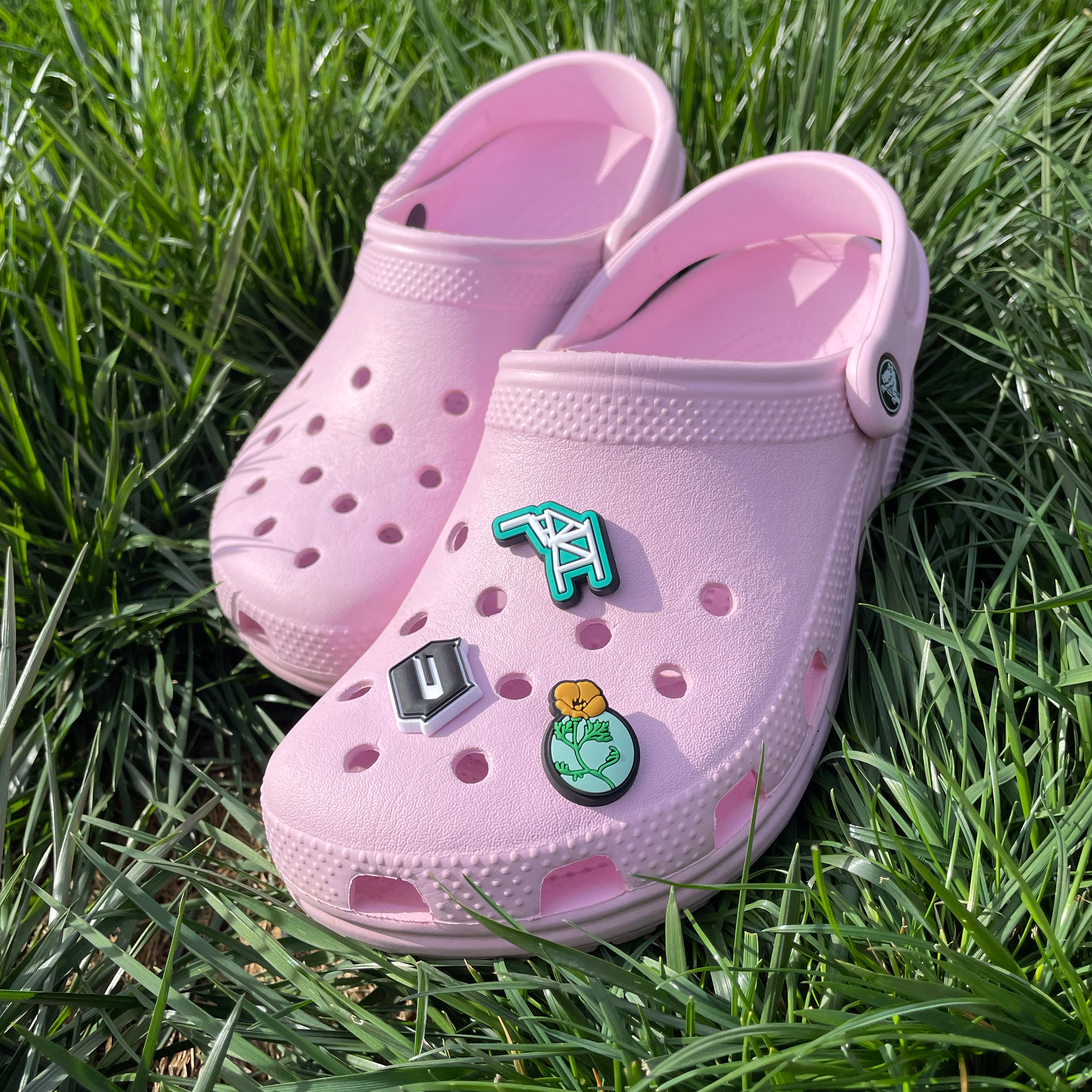 A pair of pink clogs is placed on lush green grass. The Crocs feature four Official Shoe Charms by Oaklandish: a green and white geometric design, a hexagonal gray charm, a crossed-out "U" symbol in black and white, and an illustration of a yellow flower with leaves in green.