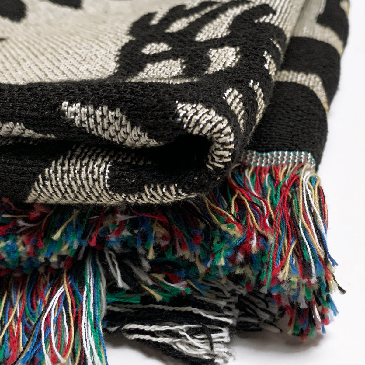 A close-up image of the folded Viva Oakland Throw by Oaklandish, featuring a distinctive black and white pattern. The edges of this cotton blanket are adorned with vibrant, multicolored fringes in shades of red, blue, green, yellow, and white. The texture and intricate weave of the fabric are clearly visible.