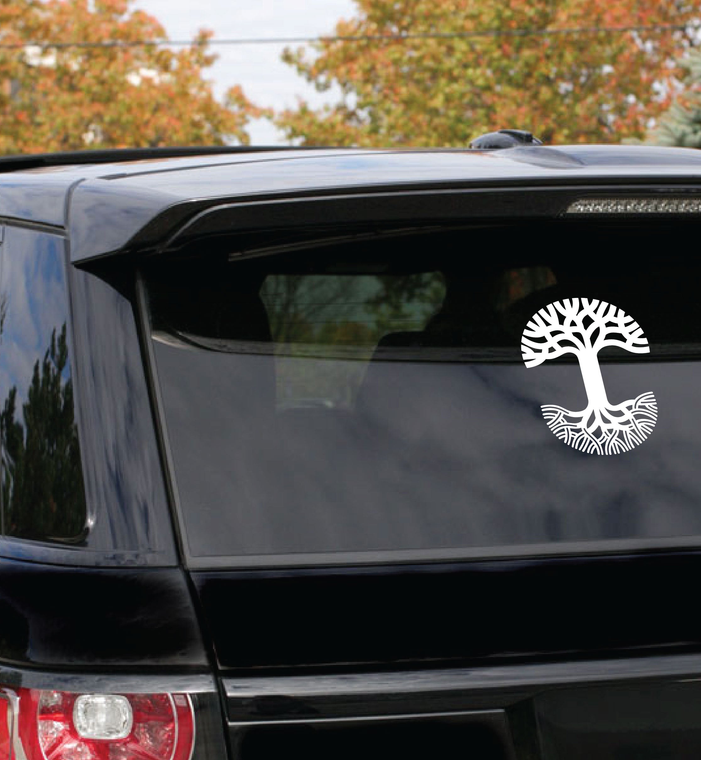 An outdoor setting in Oakland features a black SUV parked among trees adorned with autumn foliage. On the rear window of the SUV is an Oaklandish Decal 8x10 by Oaklandish, showcasing a white tree design with extensive roots forming a circular pattern and branches spreading upwards to cover the top half.