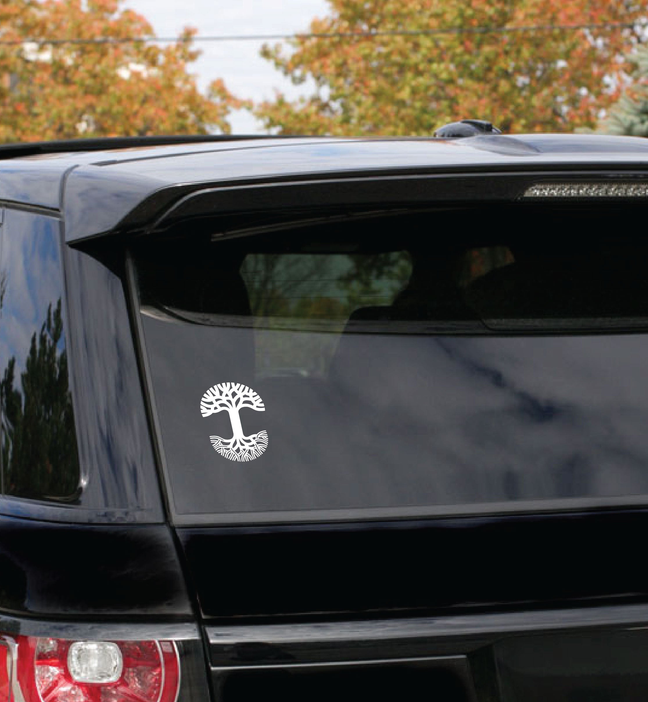 A durable black SUV with an Oaklandish Decal 5x7 white tree sticker on the rear window is parked outdoors. In the background, trees with autumn foliage are visible under a partly cloudy sky.