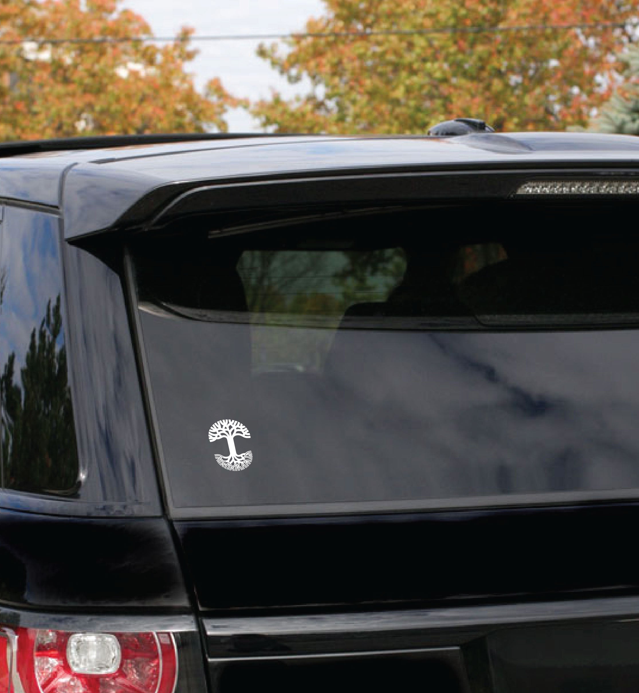 A black SUV is parked with its rear window visible, adorned with a small white Oaklandish Decal 3X4.5 depicting the Tree of Gondor. The trees in the background suggest an autumn setting, with their leaves showing various shades of orange and green. The vehicle is partially in shade, reflecting clear autumn light on its durable outdoors finish.